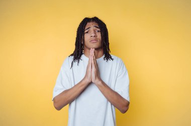 Saçları örgülü genç bir adamın portresi. Sarı arka planda dua pozisyonunda pozlar veriyor.