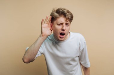 Dikkatle dinlerken şaşkınlığını ifade eden genç bir adamın portresi, bej arka planda merak ve nişan sergiliyor.