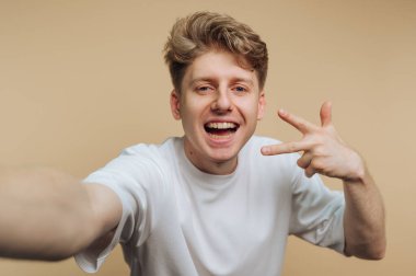 Parlak bir gülümseme ve neşeli barış işareti jesti ile bej arka planda pozitiflik ve neşe saçan genç bir adamın portresi