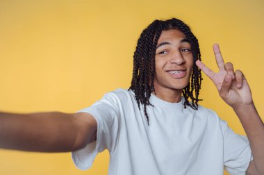 Genç bir adamın mutlu bir şekilde selfie çekerken bir barış işareti göstermesi, sarı arka planda mutluluğun ve kaygısız ruhun vücut bulmuş hali.