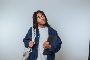 Örgülü genç bir çocuğun portresi dizüstü bilgisayar tutuyor. Beyaz arka plana bakarken düşünceli görünüyor.