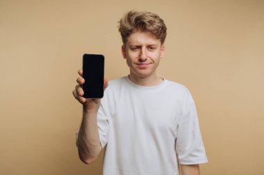 Genç bir adamın portresi, elinde akıllı bir telefon tutarken neşeli bir şekilde göz kırpar, parlak kalpli bir ruhu ve bej arka plandaki modern etkileşimi somutlaştırır.