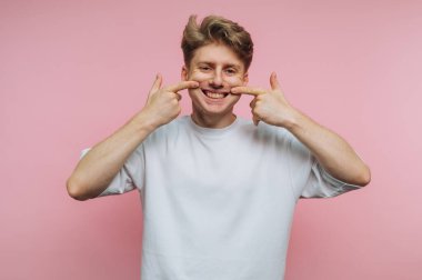 Genç oyuncu adamın portresi geniş bir şekilde gülümsüyor, şakacı bir şekilde ağzını işaret ediyor, pembe arka planda neşeli bir anı vurguluyor.