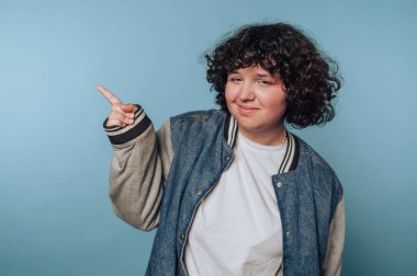 Genç, neşeli, kıvırcık saçlı bir kadının portresi mavi arka plandaki günlük kıyafetleri işaret ediyor.