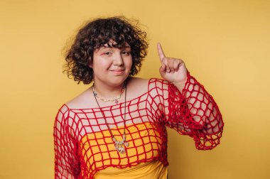 Genç bir kadının portresi göz alıcı bir kıyafetle cazibesini ve oyunculuğunu sergiliyor, sarı arka planda parmağını dudaklarına değdiriyor.