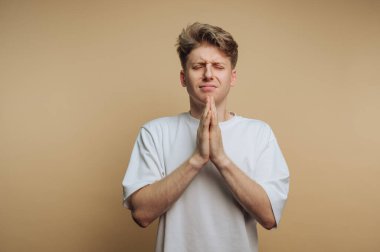 Beyaz gömlekli genç adamın portresi bej arka planda derin duyguları ifade eden bir dua jesti gösteriyor.