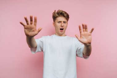 Dehşete düşmüş genç bir adamın portresi şok ve güvensizlik içinde ellerini kaldırarak pembe arka planda yoğun bir duygusal an yakalıyor.