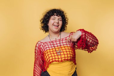 Kıvırcık saçlı, neşeli genç bir kadının portresi parlak kırmızı bir üst ve sarı arka planda sarı bir gömlek ile neşe içinde poz veriyor.