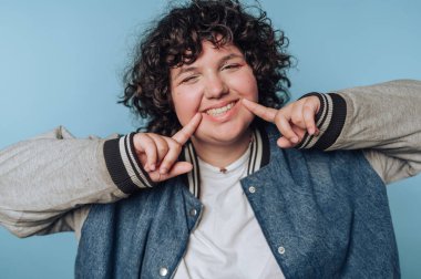 Genç ve neşeli bir kadının portresi parlak bir şekilde gülümsüyor, mavi arka plandaki kot ceketle saf mutluluk ve pozitifliği gösteriyor.