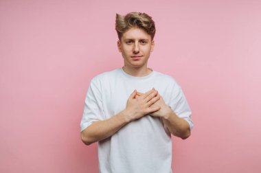Genç sakin adamın portresi ellerini nazikçe göğsüne koyuyor. Pembe arka planda sükunet ve öz yansıma sergiliyor.