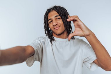 Elleriyle bir kalp yaparken gülümseyen genç bir adamın portresi beyaz arka planda sevgi dolu ve dostane bir titreşim sergiliyor.