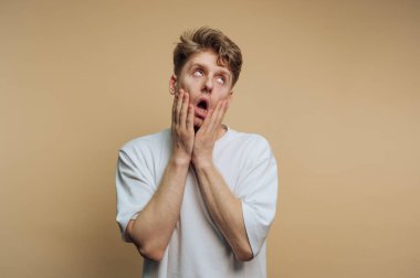 Genç adamın bej arka planda elleri üzerinde abartılı bir şok ifadesi gösteren portresi.