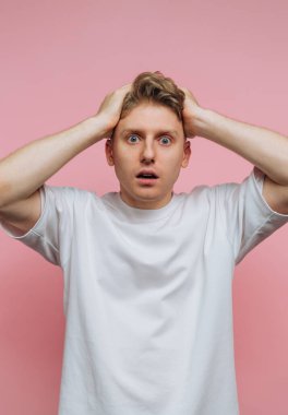 Şaşırmış genç adamın portresi kafasını tutuyor, pembe arka planda canlı bir şok ve güvensizlik tepkisi sergiliyor.