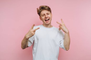 Genç ve neşeli bir adamın portresi bir gülümsemeyle barış işareti sergiliyor, pembe arka planda eğlence ve kaygısız bir ruh taşıyor.