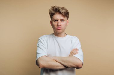 Çapraz kollar ve ciddi bir ifadeyle duran genç adam portresi bej arka planda hayal kırıklığı ve tatminsizlik gösteriyor.
