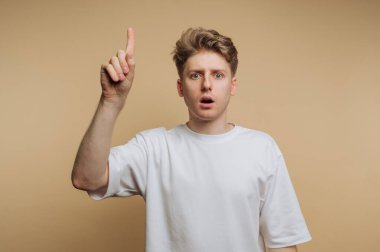 Şaşırmış bir ifade ve kaldırılmış bir parmak ile genç bir adamın portresi, bej arka planda bir fark anı veya ilham anlamına gelir.