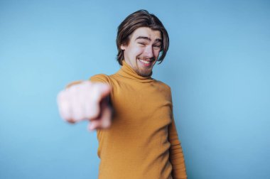 Turuncu balıkçı yakalı genç bir adamın portresi kamerayı işaret ederken gülümsüyor, mavi arka plandaki eğlenceli anları yakalıyor.