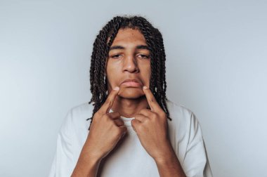 Genç bir çocuğun portresi, yüzünü çatık bir şekilde çekerek üzüntüsünü ifade eder ve beyaz arka planda duygularını göstermek için parmaklarını kullanır.