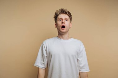 Şaşırmış genç bir adamın portresi şaşkınlıkla ağzını açmış, bej arka planda bir şok anını vurguluyor.