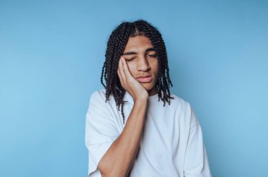 Saçları örgülü genç bir adamın portresi yüzünde yorgunluk, yorgunluğu ve mavi arka plandaki duygusal bedeli temsil ediyor.