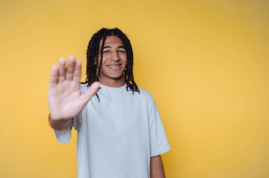 Genç adamın portresi gülümsüyor ve kamerayı selamlıyor, pozitif ve misafirperver bir atmosfer yayıyor, sarı arka planda gelişigüzel giyiniyor.