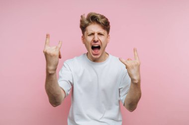 Genç dinamik adamın portresi tutkulu bir şekilde elleri ile bir kaya işareti yapıyor. Pembe arka plandaki enerjik kaya titreşimlerini gösteriyor.