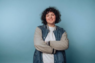 Kıvırcık saçlı, gülümseyen ve kollarını kavuşturan genç bir kadının portresi, mavi arka planda rahat bir tavır sergiliyor.