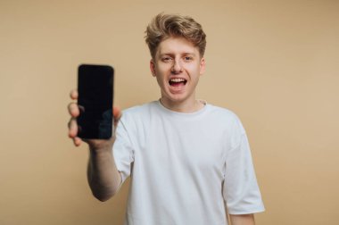 Genç adamın portresi neşeli bir şekilde gülümserken akıllı telefonunu sergiliyor, heyecan yakalıyor ve bej arka planda modern bir yaşam tarzını yakalıyor.