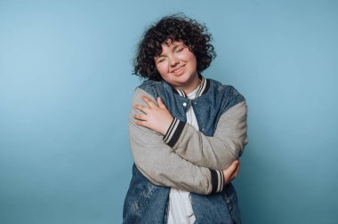 Kollarını sarıp sarmalayan genç bir kadının portresi mavi arka planda kendini sevme ve rahatlığı somutlaştırıyor.