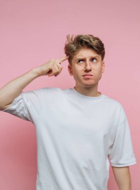 Düşünceli bir gencin portresi pembe arka planda düşünceli bir yüz ifadesi sergileyerek parmağını kafasına vuruyor.