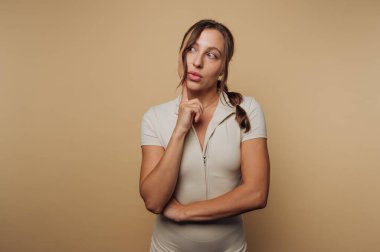 Düşünceli genç bir kadının portresi, bej arka planda bej bir elbise giymiş, parmağını çenesine dayamış düşünceli bir şekilde poz verir.