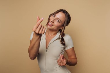 Genç, neşeli bir kadının portresi, parmaklarını kullanarak bir kalp hareketi ile poz veriyor. Bej arka planda sıcaklık ve pozitiflik yayıyor.