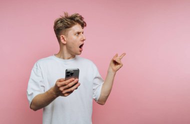 Şaşkın genç adamın portresi telefonuyla jestler yapıyor. Pembe arka planda şoku ve merakı canlı bir şekilde ifade ediyor.