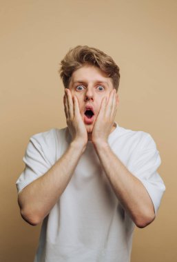 Genç bir adamın portresi, bej arka planda şaşkınlık ve şaşkınlık içinde yüzünü iki eliyle tutarak şok gösteriyor.