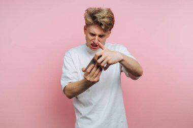 Hüsrana uğramış genç bir adamın portresi, pembe arka planda rahatsızlık ve hoşnutsuzluk gösterirken akıllı telefonunu yoğun bir şekilde inceliyor.