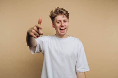 Coşku ve heyecanla işaret eden genç bir adamın portresi, bej arka planda neşe ve nişanla dolu bir anı yakalıyor.