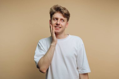 Genç bir adamın portresi, yumuşak bir gülümsemeyle duruyor, yüzüne nazikçe dokunuyor, düşünceli bir ruh hali sergiliyor, bej arka planda güzelce çerçevelenmiş.