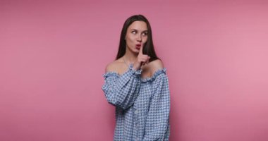 Genç bir kadının pembe arka planda mavi kareli bir elbise giyerken şakacı bir şekilde fısıldadığı ve işaret verdiği görüntüler bir sırdır.