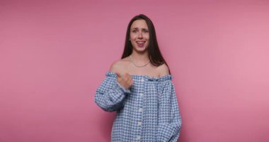 Mavi elbiseli genç bir kadının pembe arka planda baş parmağıyla poz vermesi.