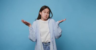 Genç bir kadının mavi arka plandaki bir durumla ilgili belirsizliği tasvir eden açık avuç içi omuz silkerek kafa karışıklığını gösteriyor.