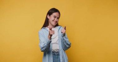 Neşeli genç bir kızın neşeli pozları başparmak hareketleriyle, coşkulu bir coşkuyla sarı arka planda pozitiflikle