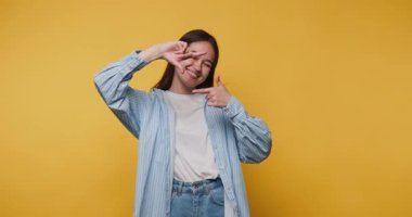 Genç ve neşeli kadının görüntüsü parmaklarıyla neşeli bir şekilde yüzünü çerçeveler, sarı arka planda neşe ve yaratıcılık saçar.