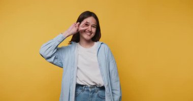 Neşeli genç bir kadının neşeli bir barış işaretiyle poz verdiği görüntüler. Sarı arka planda mutluluk ve gençlik yayıyor.