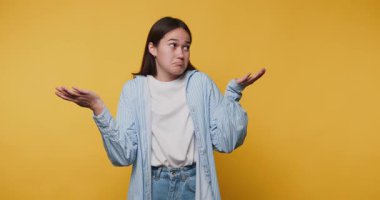 Genç bir kadının görüntüsü şaşkın ve kararsız görünüyor. Sarı arka planda ellerini omuz silkerek kaldırıyor.