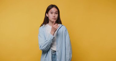 Genç bir kadının, parmaklarını dudaklarına dayadığı, gizemli bir titreşim yaydığı ve sarı arka planda sessizlik istediği görüntüler.