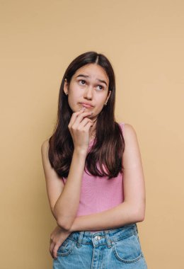 Genç bir kız parmağını dudaklarına dayayıp düşünceli bir bakışla yukarıya doğru bakan düşünceli bir şekilde duruyor. Kolsuz pembe bir bluz ve kot şort giyiyor. Etrafı bej rengi bir arka planla çevrili..