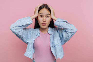 Genç bir kadın parlak pembe bir arka planın önünde duruyor, görünür bir şekilde şaşırmış ve şok olmuş. İki eliyle başını tutup beklenmedik bir duruma karşı güçlü bir duygusal tepki veriyor..