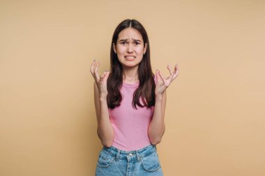 Genç bir kadın, sıkıntı ve hüsran belirtileri gösteren düz bir arkaplanda duruyor. Öfkeden ellerini kaldırıyor pembe bir bluz ve kot pantolon giyiyor, duygularıyla boğulmuş bir halde..