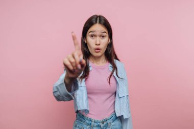 Genç bir kadın pembe bir arka plana karşı duruyor ve anlamlı bir jest yaparak işaret parmağını kaldırıyor. Şaşırmış yüz ifadesi bir vurgu anını ya da önemli bir noktayı işaret ediyor..
