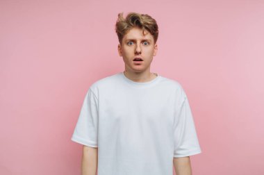 Kısa, dağınık saçlı genç bir adam pembe yumuşak bir duvara karşı duruyor ve şaşkınlık ifadesi sergiliyor. Düz beyaz bir gömlek giyiyor ve sıradan bir iç mekanda gibi görünüyor..